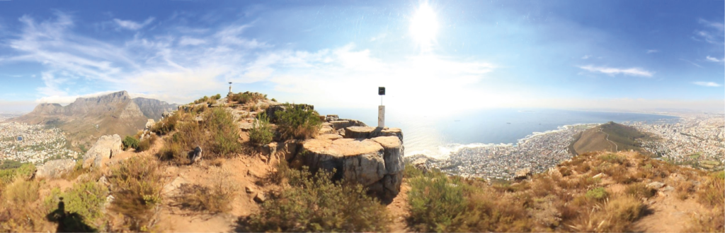 Panorama am Ende des Gipfel-Plateaus mit kleinen Büschen, flachen staubigen Stellen und flachen Felsen zur Rast, sowie mit weitem Blick über Kapstadt, rechts Signal Hill und der Ozean, und links im Hintergrund der Tafelberg und die Zwölf Apostel
