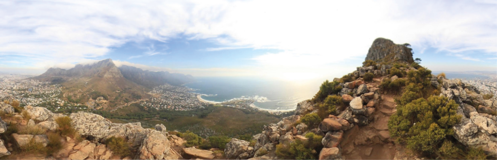 Panorama während des Umwegs, schmaler steiniger und staubiger Weg gesäumt von kleinen Büschen, rechts im Hintergrund die Spitze des Lion's Head, mittig das Meer und der Strand von Camps Bay und links der Tafelberg wie so oft typisch eingedeckt von einem dicken Wolkenschleier