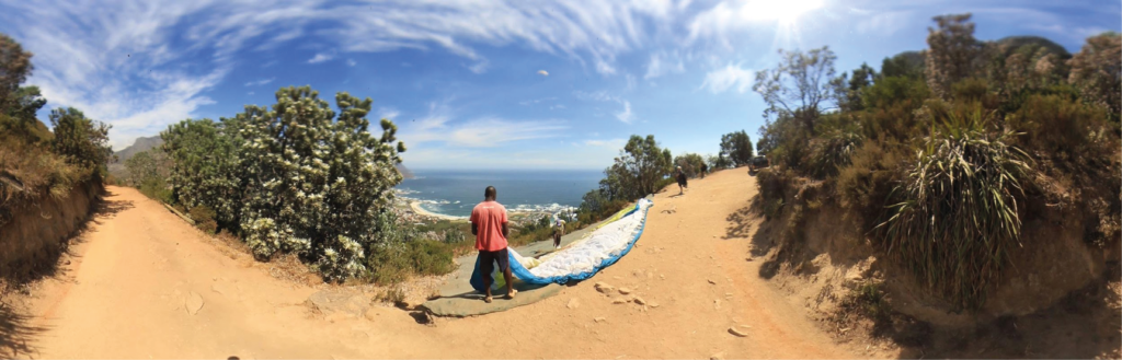 Startplatz für Paragleiter gegen Ende des ersten Viertels des Wanderwegs. Ein Helfer hält im Vordergrund den auf dem Boden ausgelegten Gleit-Schirm, hinter dem steil abschüssigen Starthang zwischen hohen Büschen: das Meer und der weite Strand von Camps Bay