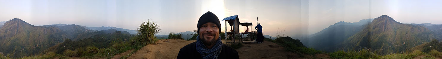 Selfie bei Sonnenaufgang mit rundum Panorama auf dem kleinen Gipfel-Plateau des Little Adam's Peak in Sri Lanka, im Hintergrund zwei kopfhohe Buddha-Statuen im Lotussitz unter kleinen Dächern sowie weitere in Morgendunst getauchte Berggipfel