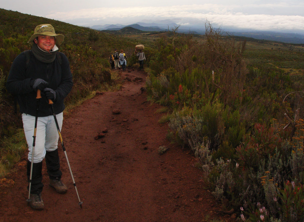 Foto beim Abstieg eines rostbraunen, lehmigen, stärker bergab führenden Wegabschnitts auf dem Kilimandscharo etwa auf der dritten Etappe der Marangu Route, eingerahmt in satten grünen mit vereinzelt rötlich blühenden Gebirgssträuchern sowie einem weiten Blick bergab auf Wolken und kleinere Berge in der Ferne – links im Vordergrund steht der Verantwortliche für HikingTrailView.com mit Wanderstöcken, mit Lehm verschmierten Bergstiefeln, kniehohen Gamaschen, dünnen Handschuhen, einem grünen Mount-Kilimanjaro-Hut mit breiter Krempe sowie sonnenverbranntem Lächeln, mittig im Hintergrund die Rücken anderer Wanderer beim Abstieg sowie ein Träger, der uns mit großem Gepäcksack auf dem Kopf beim Aufstieg entgegenkommt