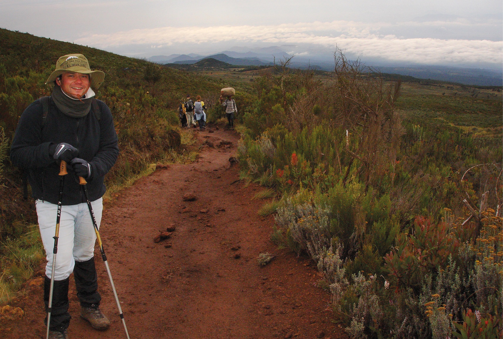 Foto beim Abstieg eines rostbraunen, lehmigen, stärker bergab führenden Wegabschnitts auf dem Kilimandscharo etwa auf der dritten Etappe der Marangu Route, eingerahmt in satten grünen mit vereinzelt rötlich blühenden Gebirgssträuchern sowie einem weiten Blick bergab auf Wolken und kleinere Berge in der Ferne – links im Vordergrund steht der Verantwortliche für HikingTrailView.com mit Wanderstöcken, mit Lehm verschmierten Bergstiefeln, kniehohen Gamaschen, dünnen Handschuhen, einem grünen Mount-Kilimanjaro-Hut mit breiter Krempe sowie sonnenverbranntem Lächeln, mittig im Hintergrund die Rücken anderer Wanderer beim Abstieg sowie ein Träger, der uns mit großem Gepäcksack auf dem Kopf beim Aufstieg entgegenkommt
